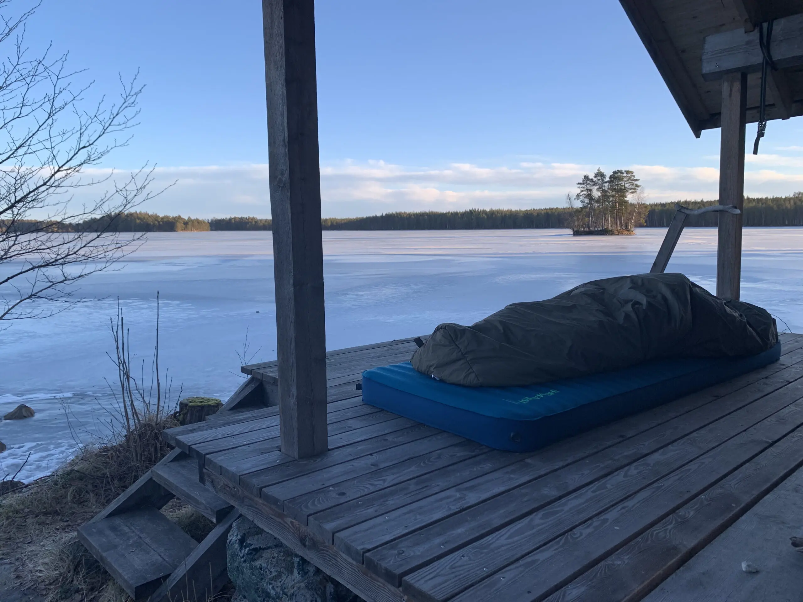 Sovepose og liggeunderlag på træterrasse ved frossen sø i naturen under en klar blå himmel.